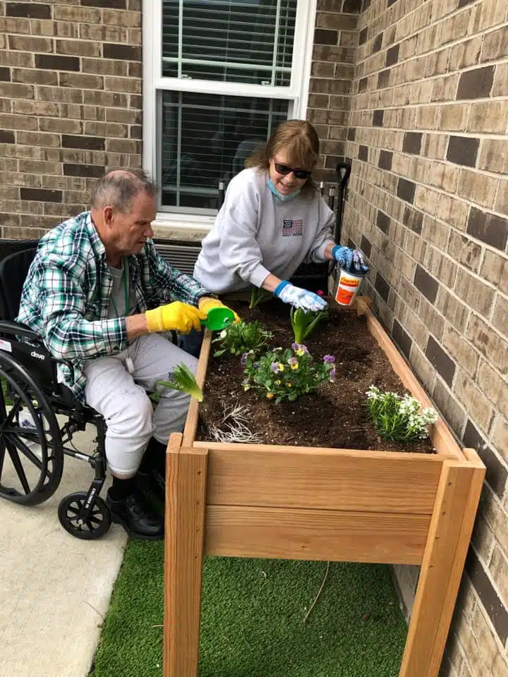 Resident in a wheelchair and caregiver planting flowers together in a raised garden bed outside the home.