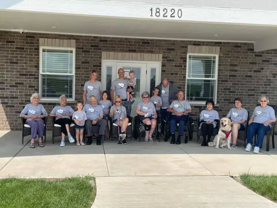 Residents, caregivers, and family members sitting together outside the assisted living home, with a service dog in front.