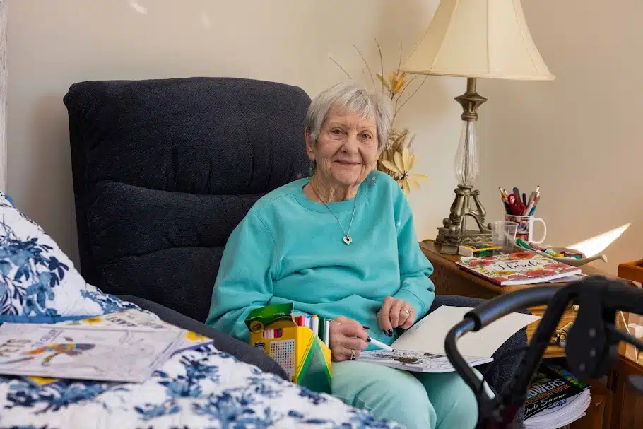 Senior woman sitting in a cozy assisted living room with coloring books, personal items, and a walker nearby while preparing for a move.